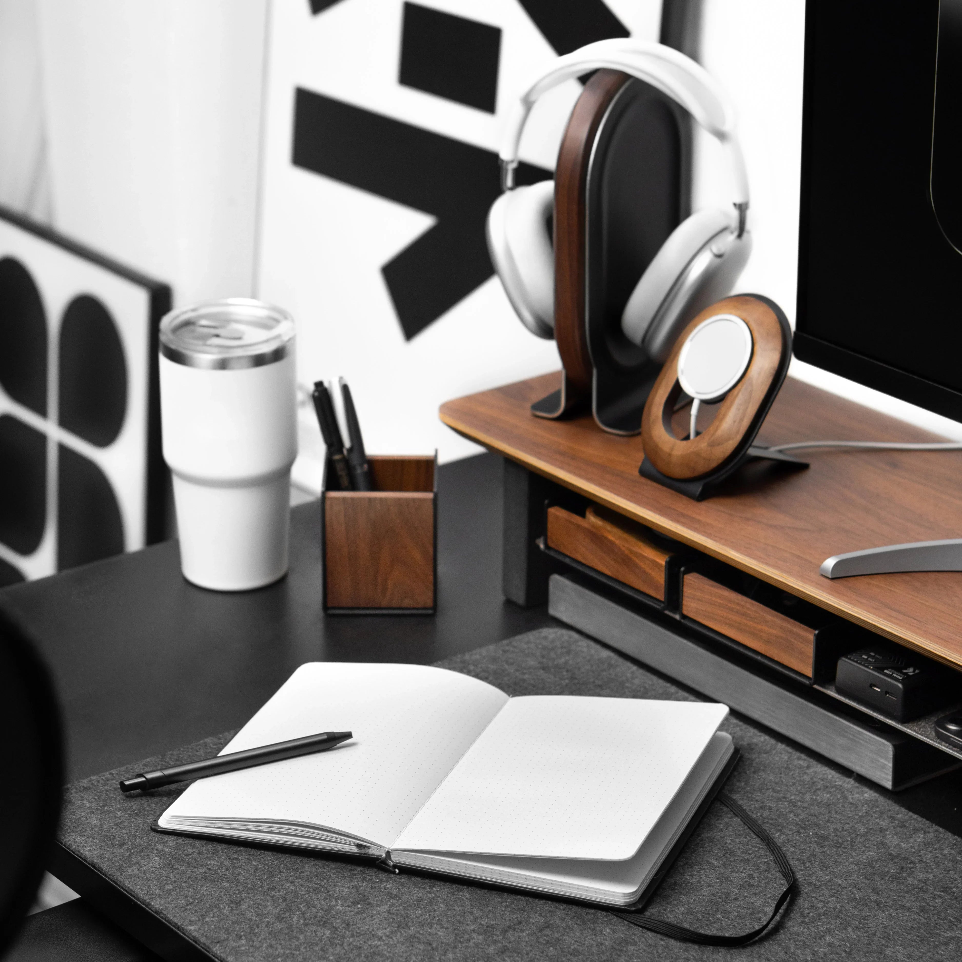 Desk setup with notebook, pen, tumbler, and headphones on a wooden stand.